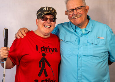 photo of Andrew Crane at the NFB National Convention 2024, holding his cane & wearing a t-shirt that says "I drive a stick," standing beside his father Dave